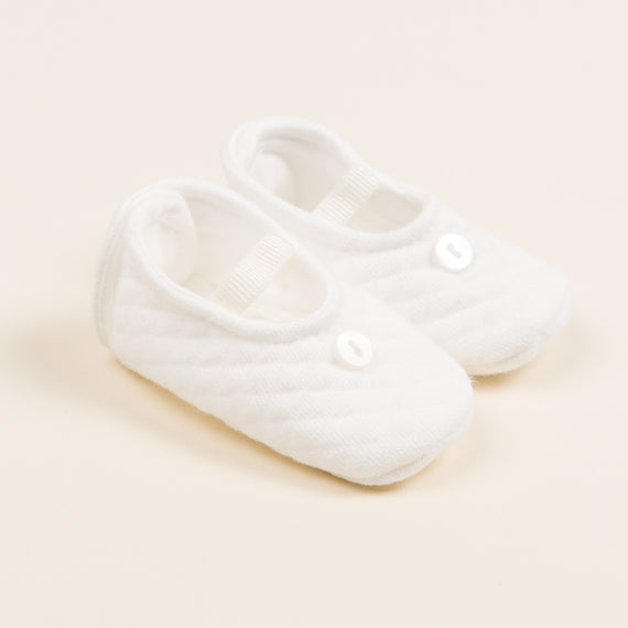 A pair of soft Ivory Quilted Booties crafted from textured cotton, featuring a small white button on each shoe. These handmade baby shoes include elastic straps over the top, all set against a light beige background.