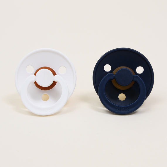 Two Bibs Pacifiers, one white and one deep space blue, placed side by side on a light beige background, ideal as an upscale baby gift.