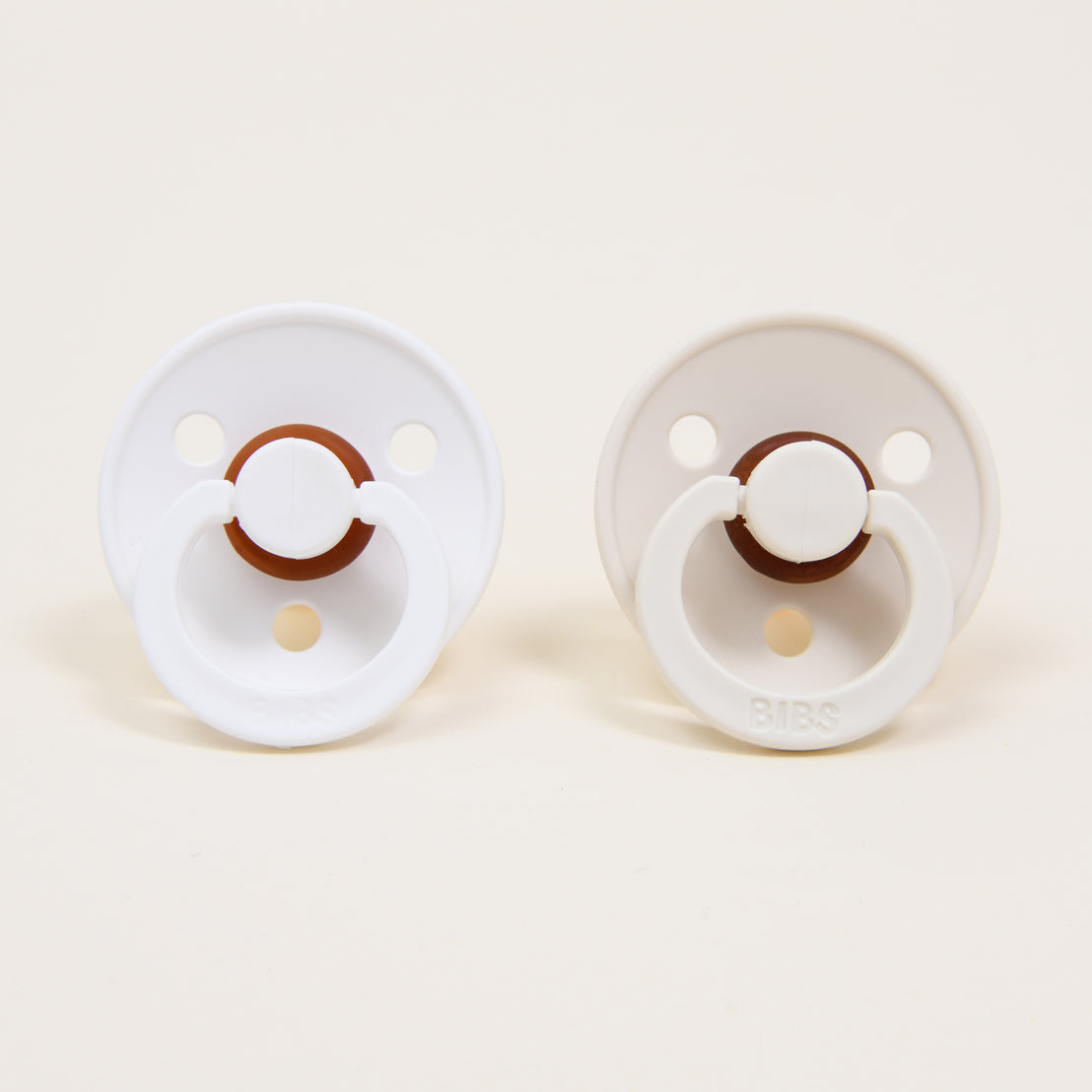 Two Bibs Pacifiers in White & Ivory on a light beige background.