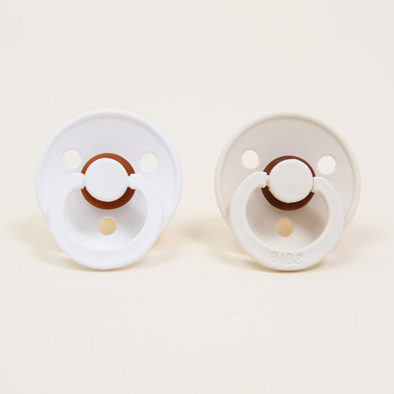 Two Bibs Pacifiers in White & Ivory on a light beige background.