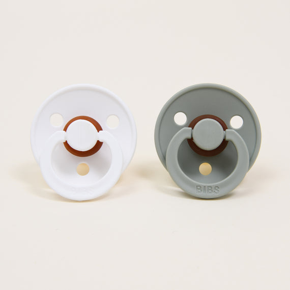 Two Bibs Pacifiers on a light background, one white and one gray, both with a button-style handle in the center branded "bibs".