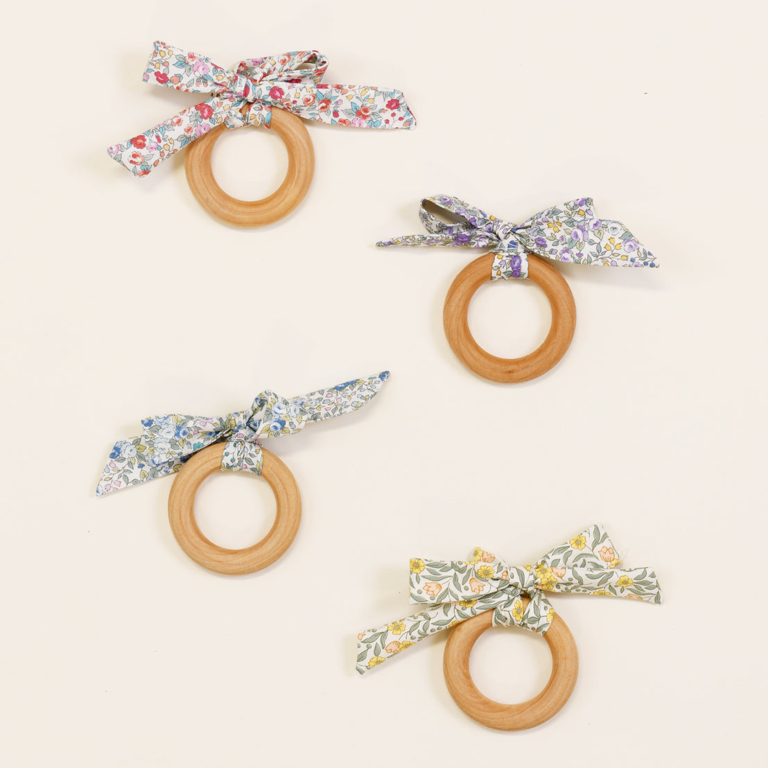 Four Petite Fleur Wooden Teether Rings, each adorned with a boutique fabric bow in different floral patterns on a plain, light background.