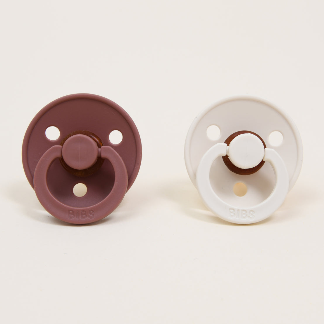 Two Bibs Pacifier Sets, one Woodchuck and one Ivory, displayed on a light background. Each upscale pacifier features a circular shape with three holes and a handle.