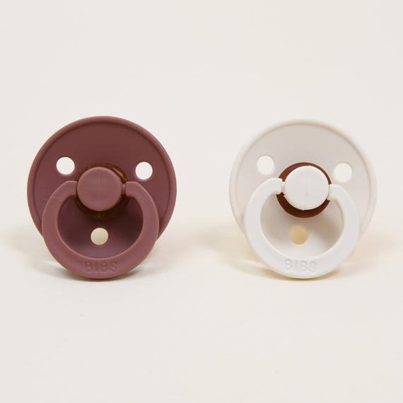 Two Bibs Pacifier Sets, one Woodchuck and one Ivory, displayed on a light background. Each upscale pacifier features a circular shape with three holes and a handle.