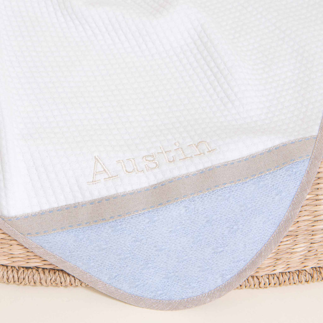 A close-up of a white and blue Austin Personalized Blanket with the name "Austin" embroidered in a delicate script, resting in a woven basket.
