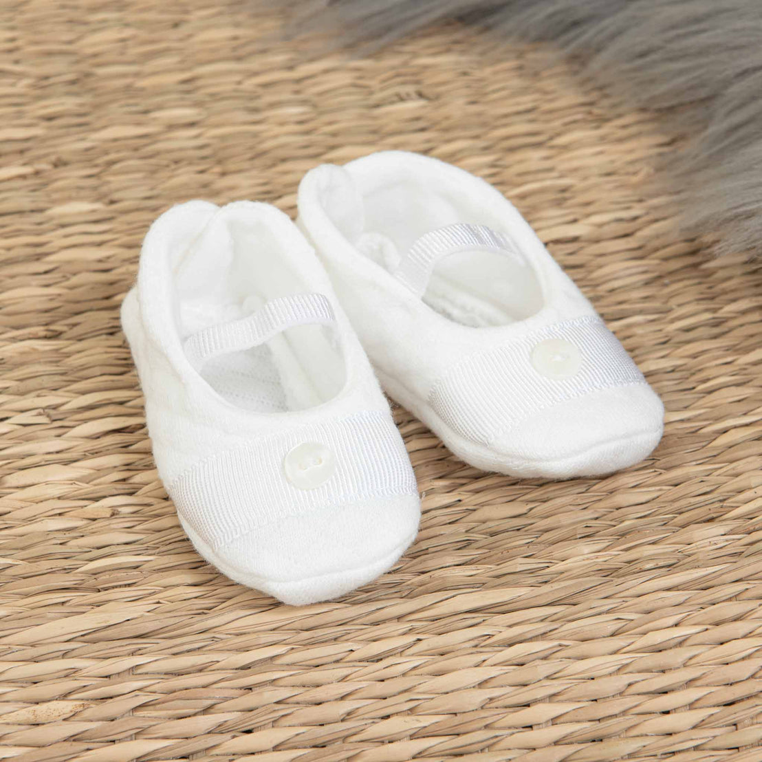 A pair of small white Elijah Newborn Booties with straps and buttons, ideal for a baptism outfit, placed on a woven textured surface. The background features a soft, gray, furry fabric.