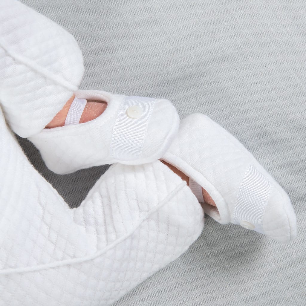 A newborn baby in a white quilted onesie lies on a light gray sheet, perfectly dressed for the occasion. Completing the charming baptism outfit with elegant snap button details, the little feet are snug in matching booties known as Elijah Newborn Booties.