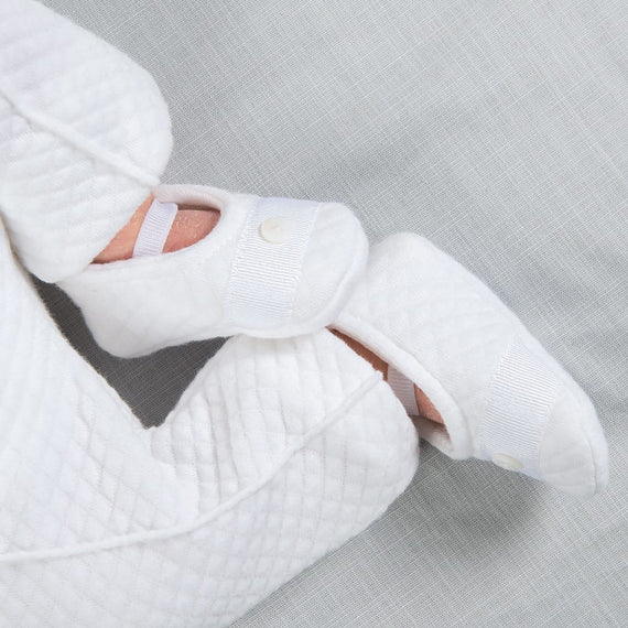 A newborn baby in a white quilted onesie lies on a light gray sheet, perfectly dressed for the occasion. Completing the charming baptism outfit with elegant snap button details, the little feet are snug in matching booties known as Elijah Newborn Booties.