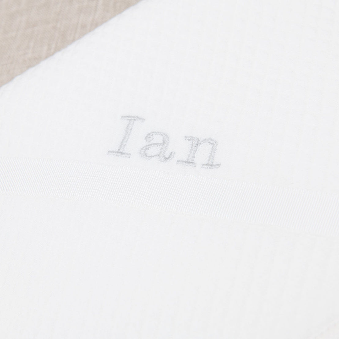 Close-up of a white fabric with the name "Ian" embroidered in gray thread, centered above a horizontal satin ribbon on the Ian Personalized Blanket in a traditional designer style.