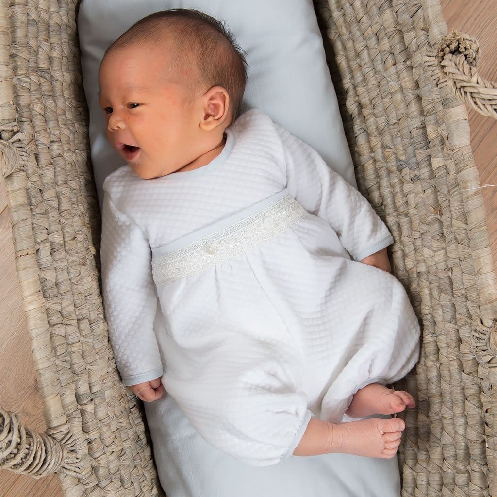 A newborn baby lying in a woven basket, dressed in the Harrison Newborn Romper with textured white fabric and charming lace accents.