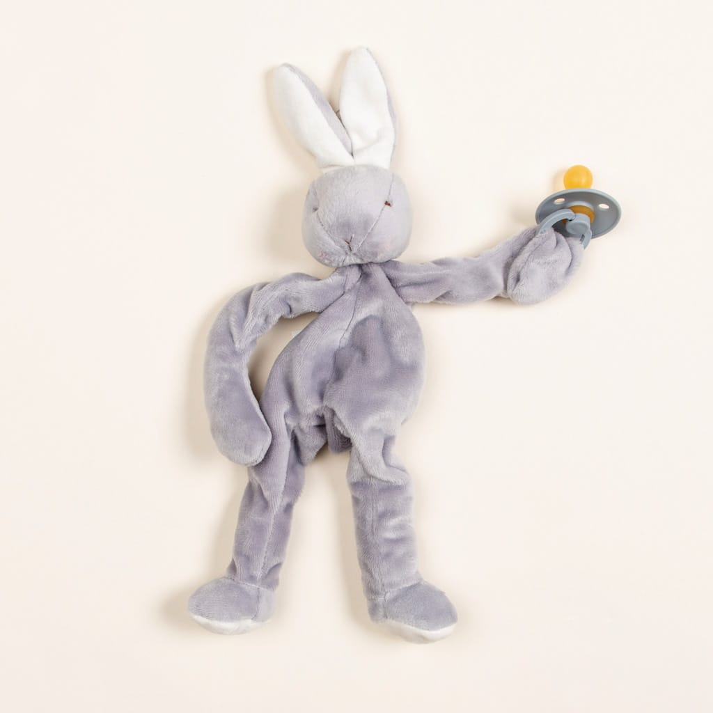 A soft, plush Grey Silly Bunny Buddy toy with long ears and a vintage pacifier attached to its paw, set against a neutral beige background.