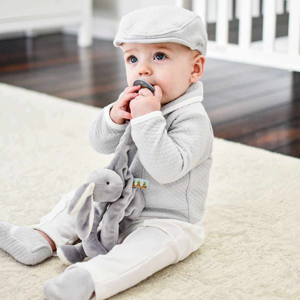 A baby wearing a Grey Silly Bunny Buddy | Pacifier Holder and sweater sitting on the floor, chewing on a stuffed elephant's ear, with a white fence in the background.