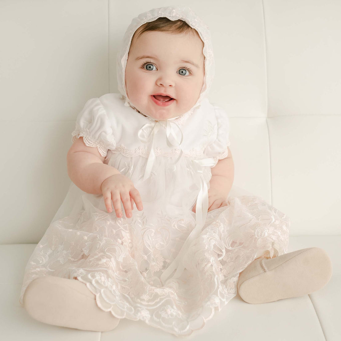 A joyful baby wearing the Joli Fitted Bonnet and matching dress sits on a white leather sofa and looks up with bright blue eyes and an open-mouthed smile.
