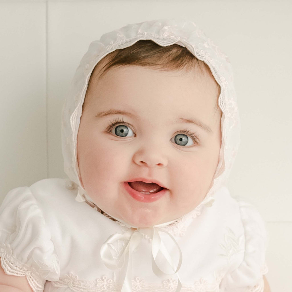 Baby girl wearing the Joli Fitted Bonnet  with lace detailing and matching dress looks upwards with big blue eyes and a joyful expression. 
