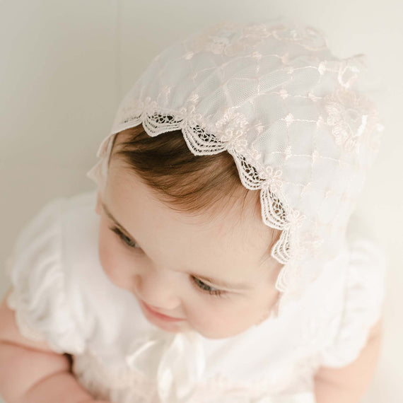Overhead view of a baby girl in the Joli Fitted Bonnet, adorned with intricate floral lace and delicate scalloped trim. The bonnet is secured with satin ribbon ties, perfectly framing her face as she gazes downward.