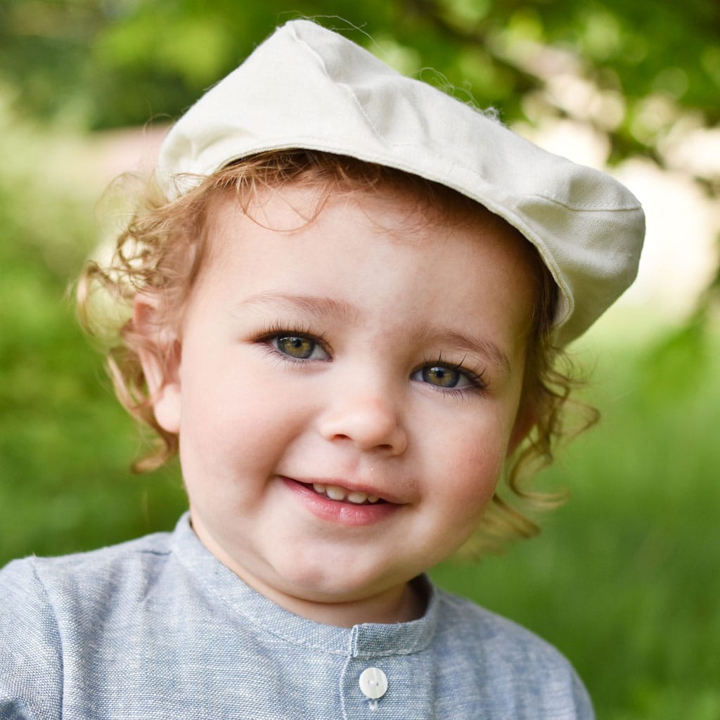 A young child, grinning in an ivory linen shirt and wearing the Ivory Linen Newsboy Cap handmade in the USA, enjoys the outdoors against a softly blurred green backdrop, suggesting a natural setting like a park or garden.
