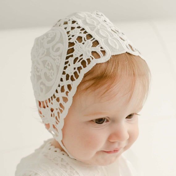A baby girl with red hair wearing a Adeline Lace Bonnet and matching lace dress looks downward away with a slight smile. 