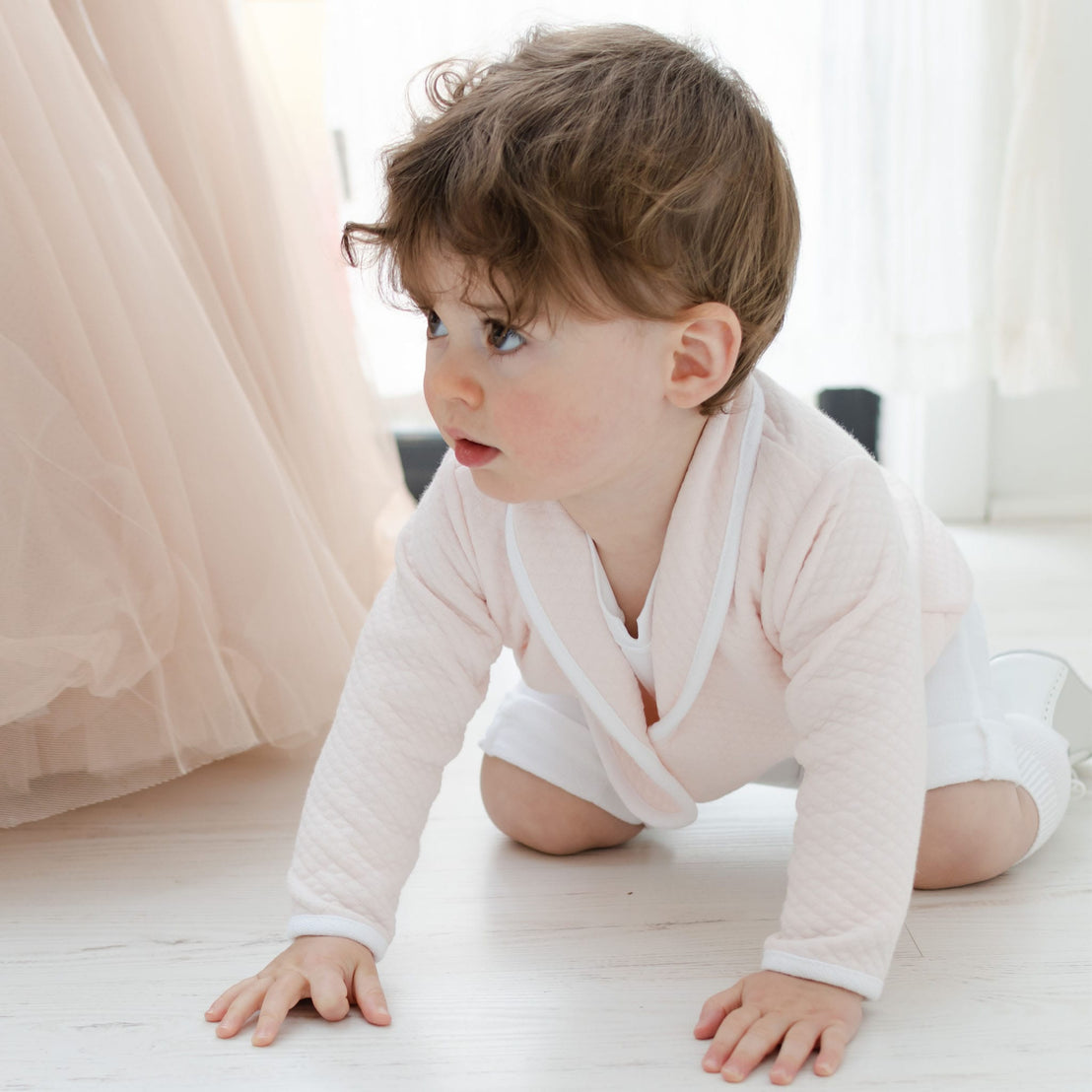 A toddler with brown hair is crawling on the floor, wearing a light pink top and the adorable Asher Blush 3-Piece Shorts Suit. The background is softly lit, with sheer curtains and part of a beige dress visible.