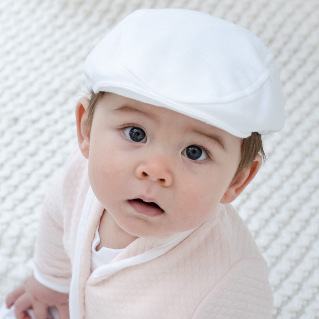 Dressed in an Asher Blush 3-Piece Shorts Suit with a matching white cap, the baby gazes up with wide eyes against the backdrop of a textured white blanket.