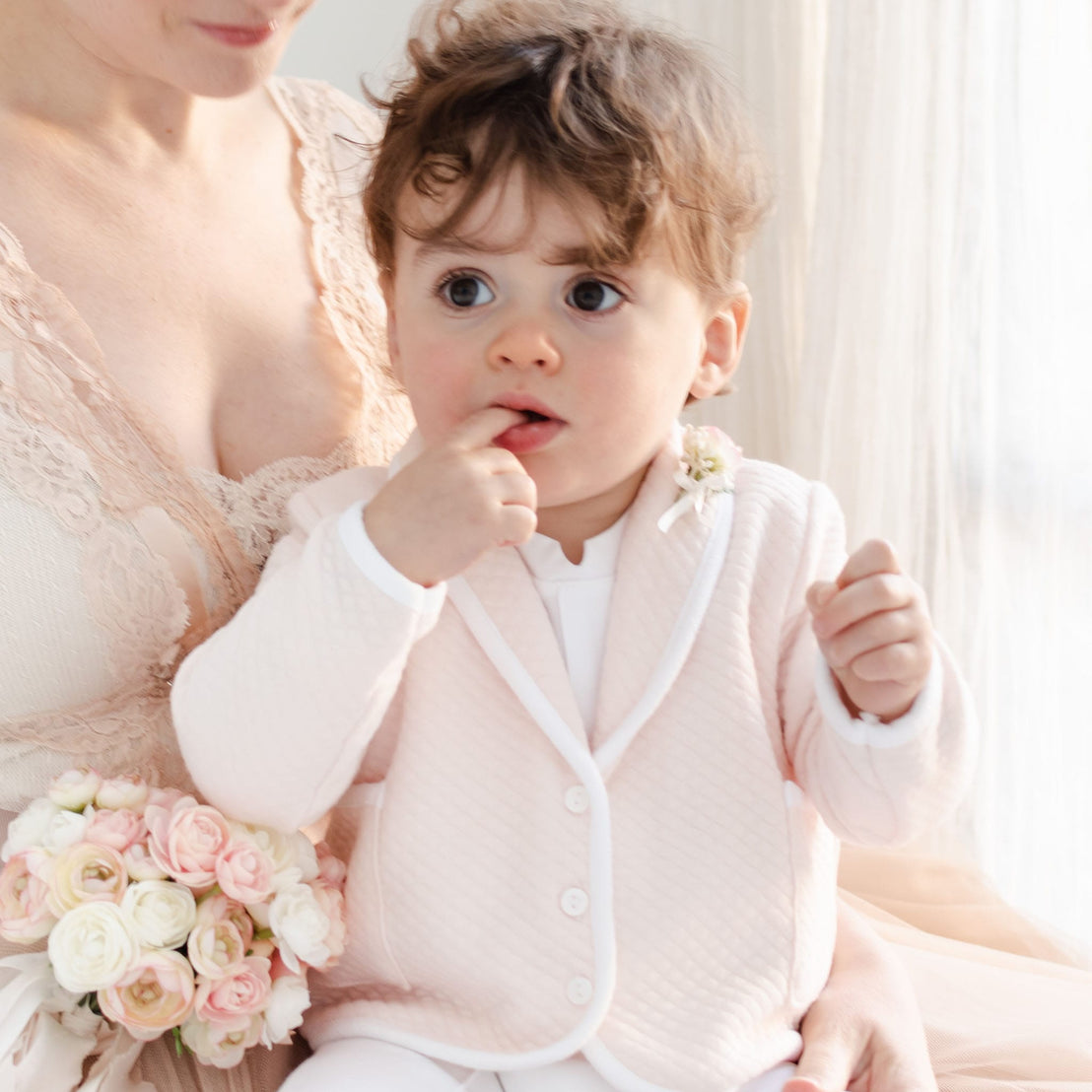 A young child with curly hair, dressed in the Asher Blush 3-Piece Shorts Suit, sits on a woman's lap. The woman, adorned in a lace dress, holds a bouquet of pastel flowers. The background features soft natural lighting filtering through sheer curtains.