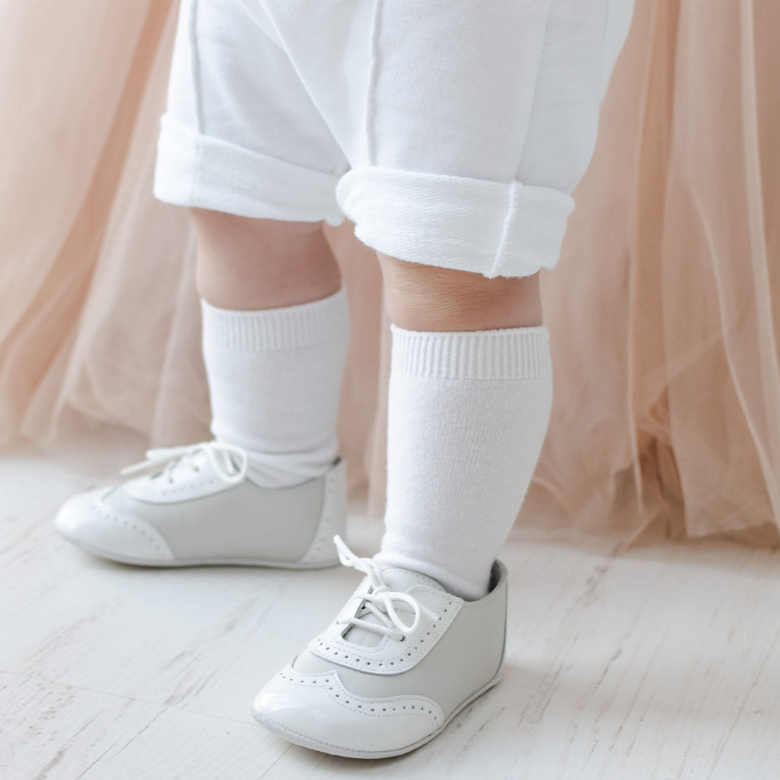 A baby wearing white knee socks and light gray lace-up shoes stands on a white wooden floor. They are dressed in the Asher Blush 3-Piece Shorts Suit, featuring cozy newborn attire with white rolled-up pants, while a soft blush quilted cotton fabric adds charm to the background.
