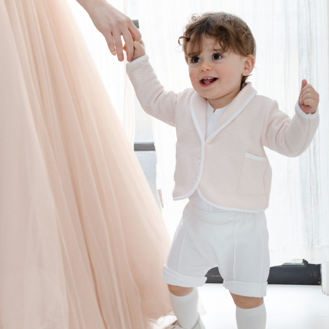 A joyful toddler wearing the Asher Blush 3-Piece Shorts Suit walks while holding an adult's hand. The adult is donned in a flowing, peach-colored dress, set against a softly lit background that highlights a cheerful, intimate moment.