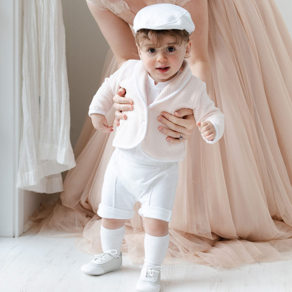 A toddler stands supported by an adult's hands, wearing the Asher Blush 3-Piece Shorts Suit, which includes a stylish white outfit reminiscent of a newborn ensemble, and light pink accents. The child pairs it with white knee socks and shoes. In the background, a soft beige flowing dress enhances the scene's elegance.
