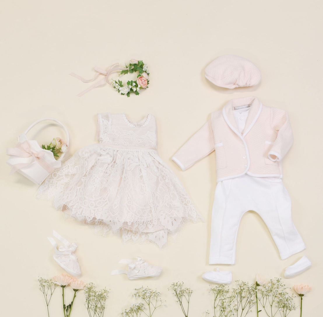 Flat lay of an exquisite Asher Blush 3-Piece Suit on a light background, featuring a lace dress, floral headband, pink jacket, white pants, coordinating hat, shoes, and a basket filled with flowers. The ensemble is arranged amid delicate small white flowers like a quilted cotton dream.