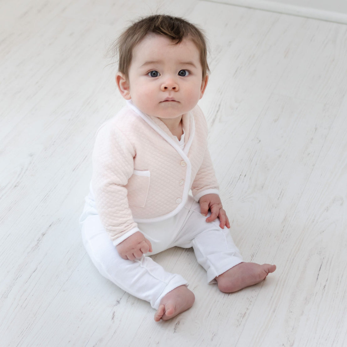 A baby with light brown hair sits on a light wooden floor, wearing the Asher Blush 3-Piece Suit. The suit includes a white baby suit and is topped with a light pink quilted cotton jacket. The baby's expression is curious and attentive, with wide eyes gazing forward, perfectly dressed for any special occasion.