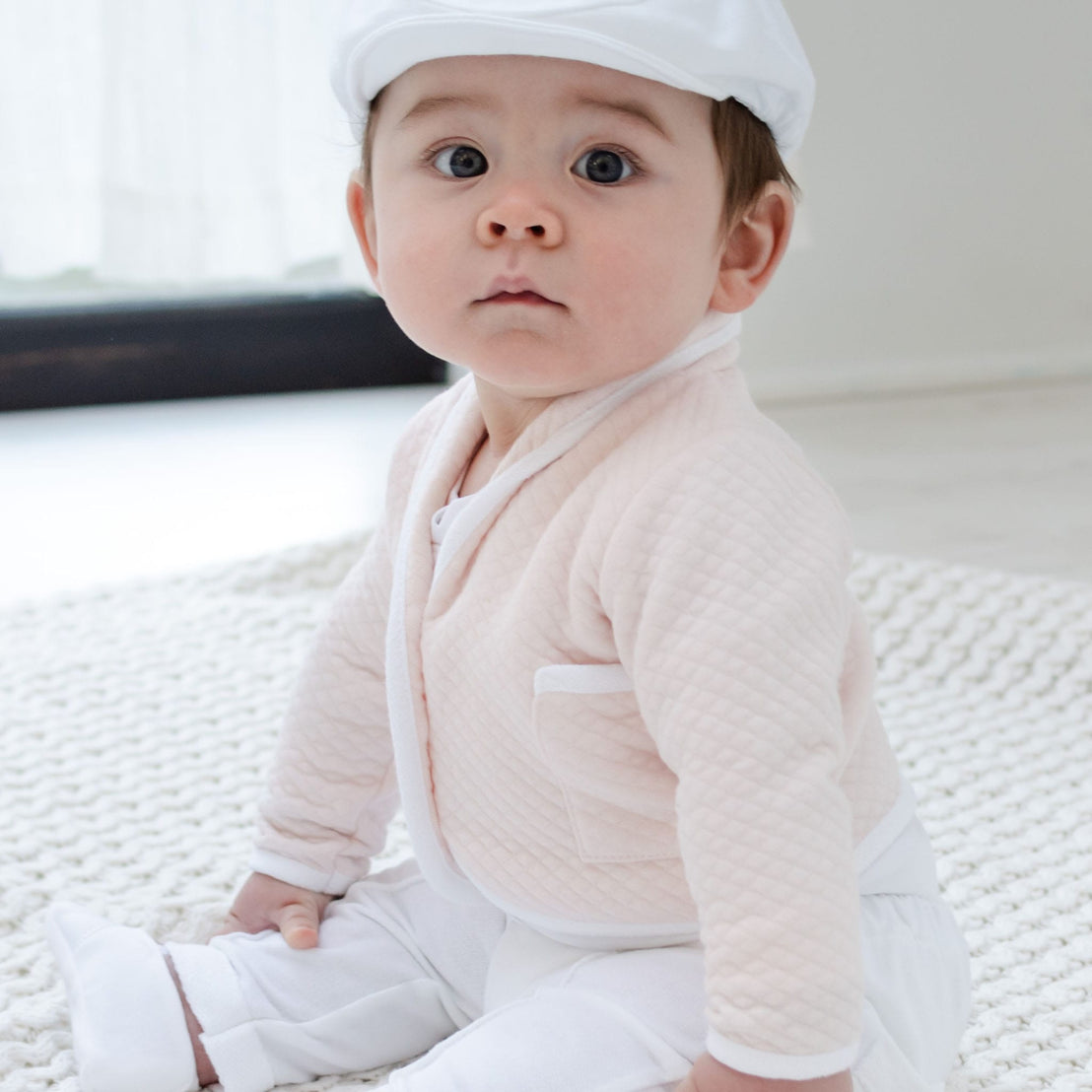 Dressed in the Asher Blush 3-Piece Suit with a delicate light pink quilted design, and paired with a white cap, a baby sits on a beautifully soft white knitted blanket. With curious eyes, the little one gazes up amidst the gentle natural light of the bright room, creating an ideal backdrop for this memorable moment.