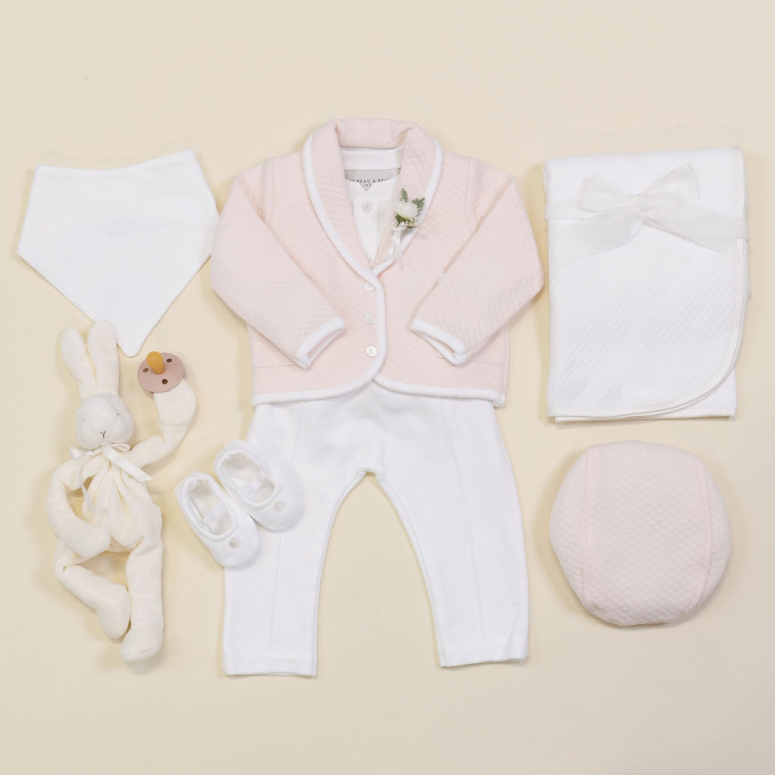 Flat lay of baby items on a neutral background showcasing the Asher Blush 3-Piece Suit, which includes a jacket and pants in pink and white tones, paired with matching shoes. The ensemble is complemented by a white bib. The setup also features a plush rabbit toy with pacifier, cozy quilted cotton blanket, and pink cushion.
