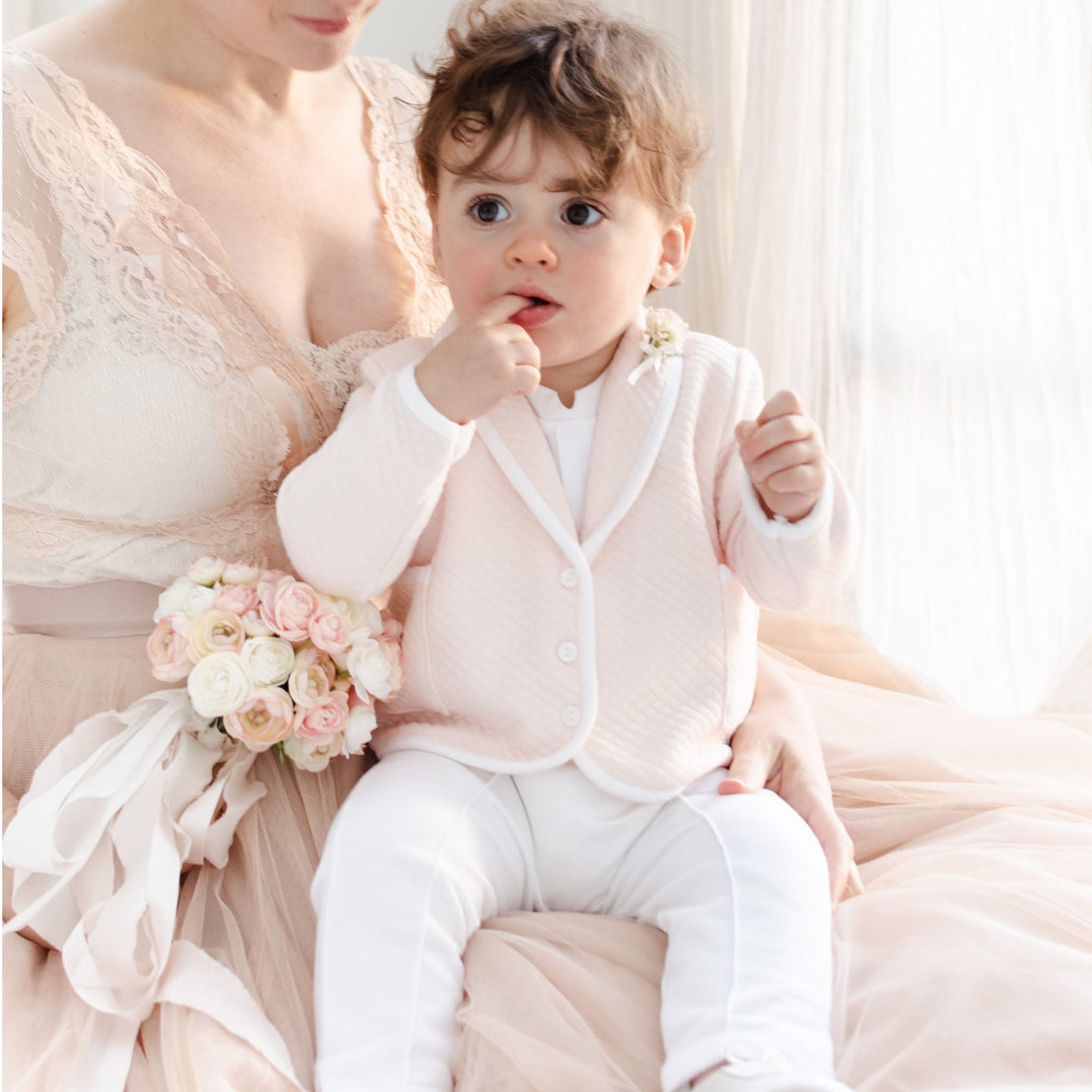 A woman in a pink lace dress holds a baby dressed in the charming Asher Blush 3-Piece Suit, which includes a light pink quilted cotton jacket and white pants. The baby, with curly hair, touches their mouth while the woman holds a delicate bouquet of white and pink roses against the softly lit background.