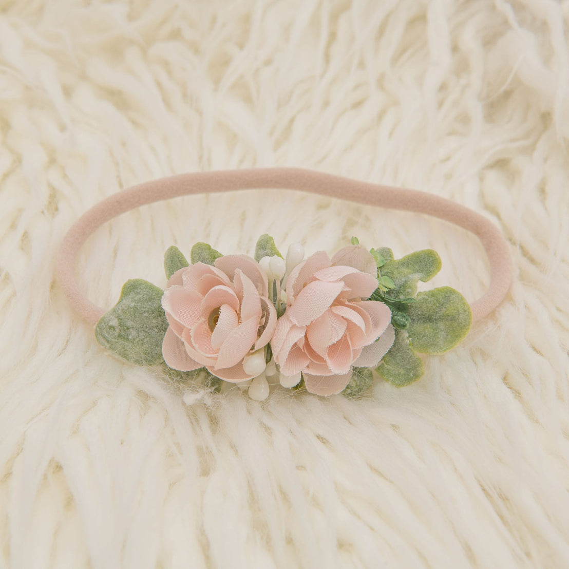 The Ava Petite Flower Headband, adorned with blush pink roses and green leaves, is displayed on a soft cream-colored surface.