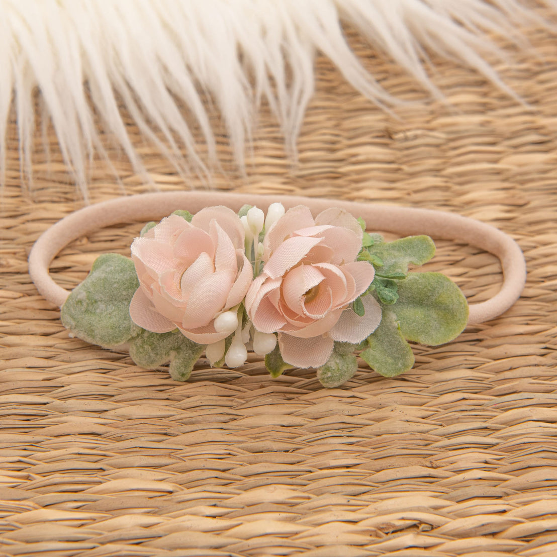 The Ava Petite Flower Headband, adorned with handmade blush pink fabric roses and green leaves, is elegantly displayed on a woven surface against a fluffy white backdrop.