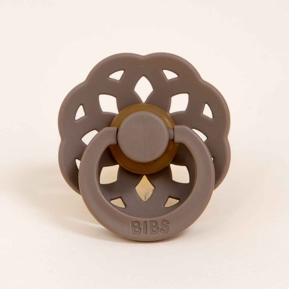 A Bibs Lace pacifier  in Dark Oak, with a circular scalloped shield and a button-style pull ring, designed and manufactured in Denmark, displayed against a plain light background.