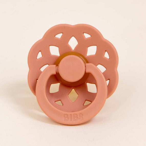 A Bibs Lace Pacifier in Peach with a bohemian inspired design, featuring a circular vented shield and a button-styled handle on a light background.