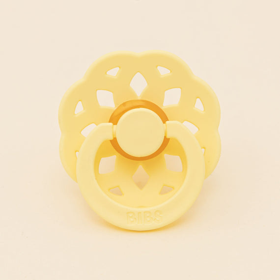 A yellow Bibs Lace pacifier with a flower-shaped shield and handle, set against a neutral background. The brand "bibs" is embossed on the front.