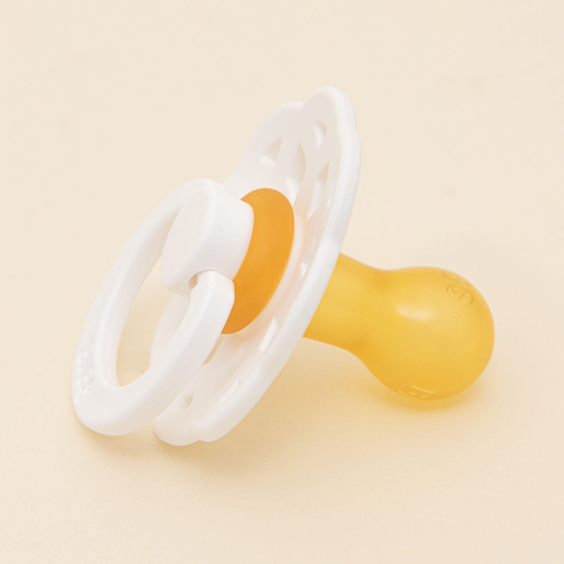 A Bibs Lace Pacifier in White with a white and transparent shield and handle, and a yellow nipple, set against a light beige background.