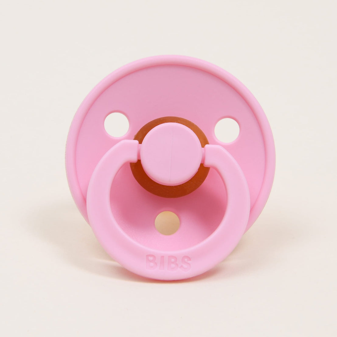 A Bibs Pacifier in Baby Pink, designed and manufactured in Denmark, with a circular rubber nipple and a round ventilation shield, labeled "bibs," on a light beige background.