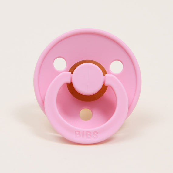 A Bibs Pacifier in Baby Pink, designed and manufactured in Denmark, with a circular rubber nipple and a round ventilation shield, labeled "bibs," on a light beige background.