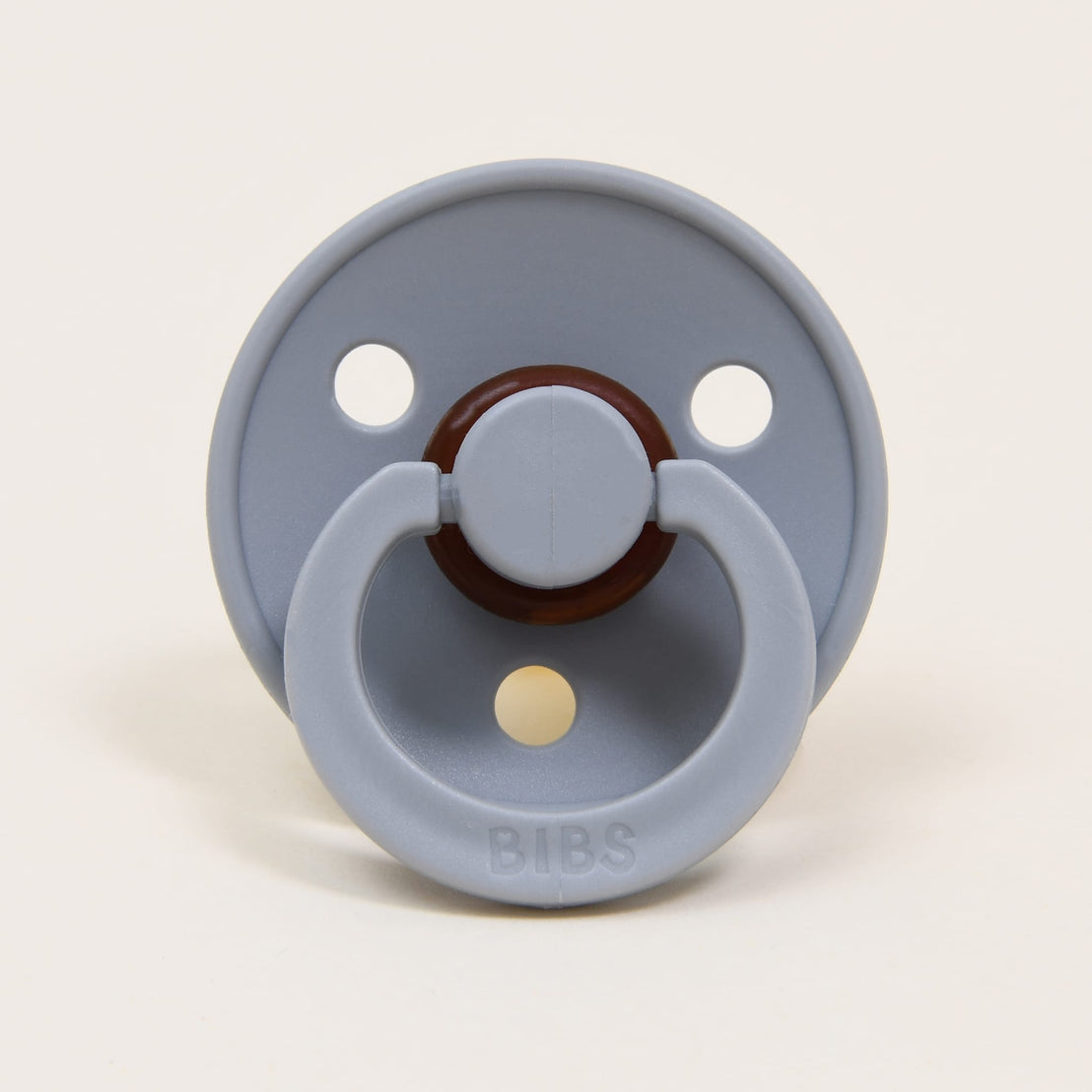A light gray Bibs Pacifier with a round shield and two air holes, featuring a brown nipple and handle, labeled "bibs" on a plain off-white background.