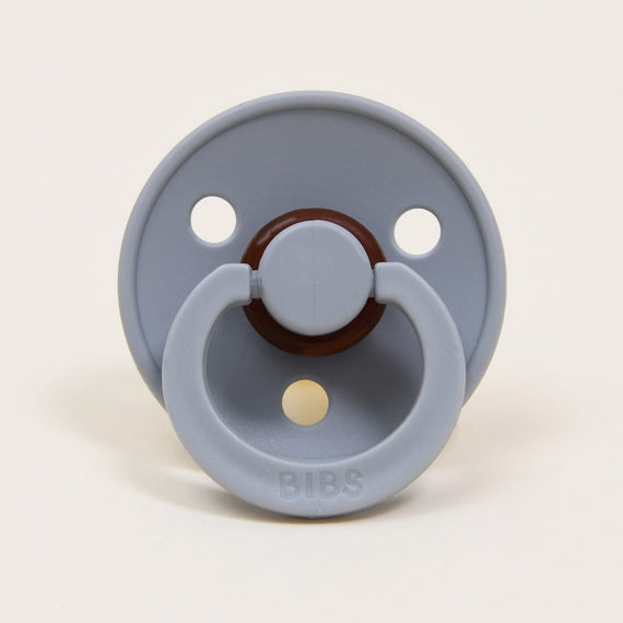 A light gray Bibs Pacifier with a round shield and two air holes, featuring a brown nipple and handle, labeled "bibs" on a plain off-white background.
