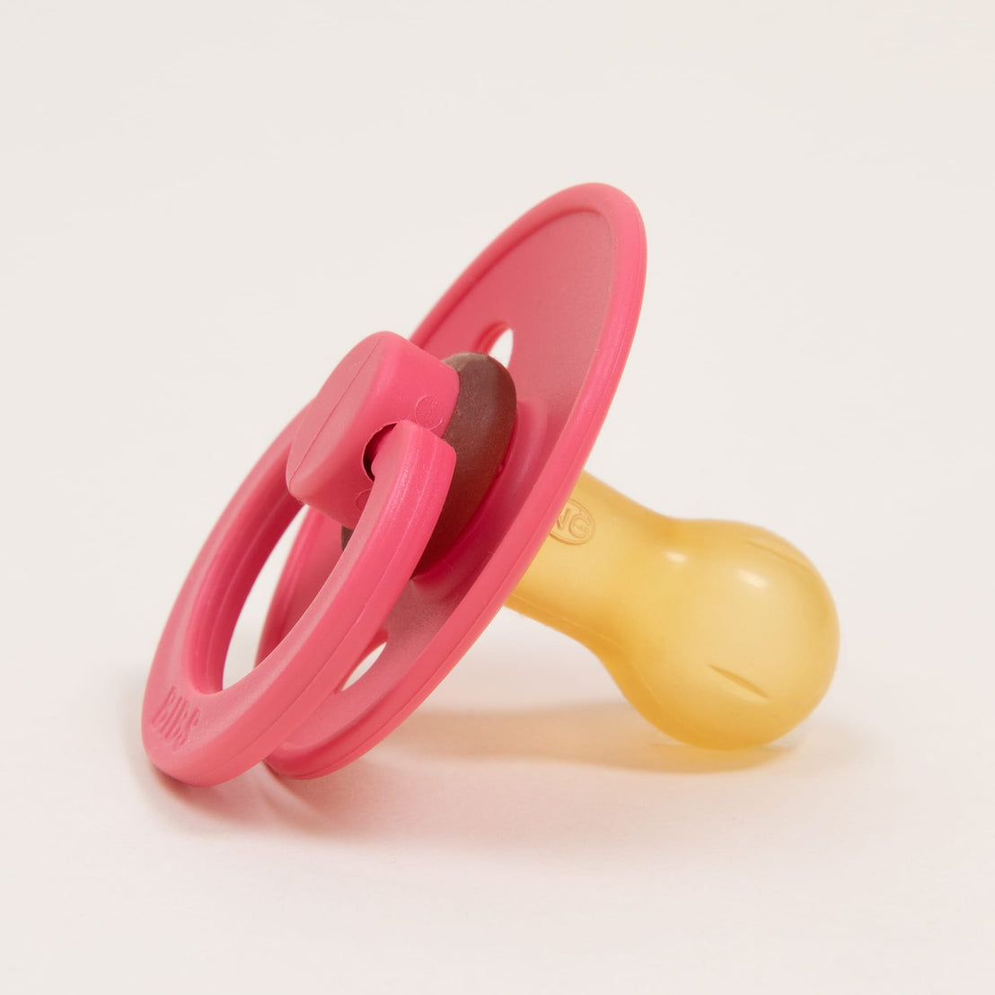 A close-up image of a pink Bibs Pacifier in Coral with a translucent yellow nipple and a circular handle on a white background.