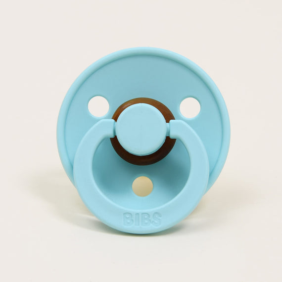 A mint green pacifier with a round brown handle and the brand name "bibs" written at the top, designed in Denmark, set against a plain white background.