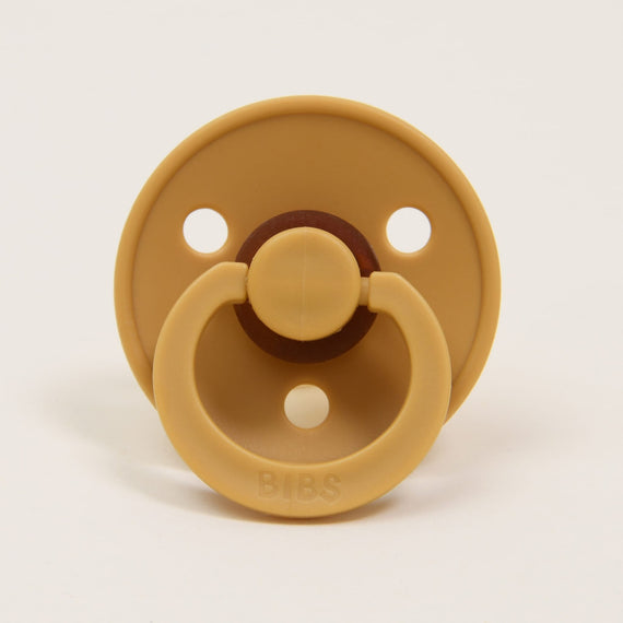 A Bibs Pacifier in Mustard yellow natural rubber baby pacifier with a circular shield and handle, along with three ventilation holes, placed against a white background.
