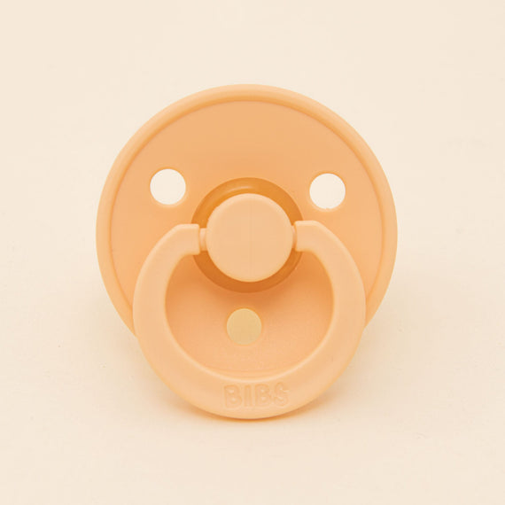 A Bibs pacifier in Peach Sunset with a round shield and two air holes, prominently featuring the brand name "bibs" on the button.