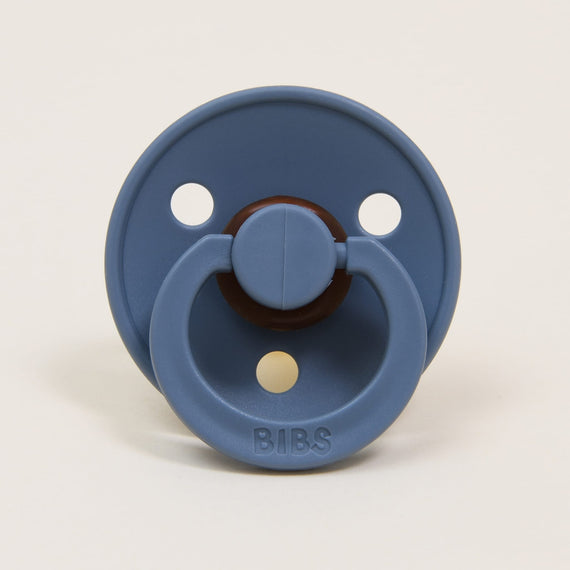 A Bibs Pacifier in Petrol that is blue with a brown handle and the word "bibs" written on it, set against a light beige background.