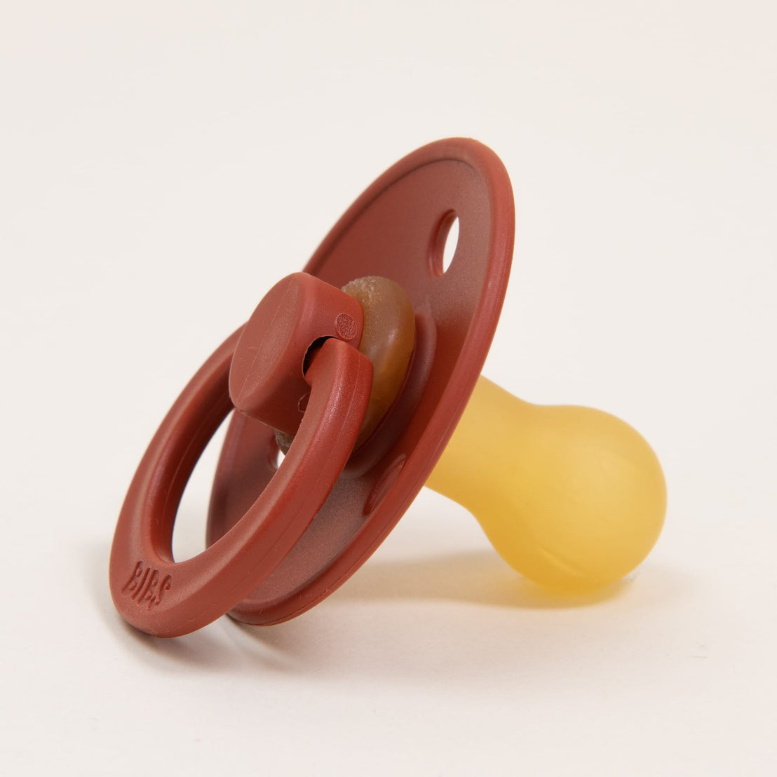 A close-up image of a Bibs Pacifier in Rust with a terracotta colored upscale and a translucent yellow nipple, on a light background.