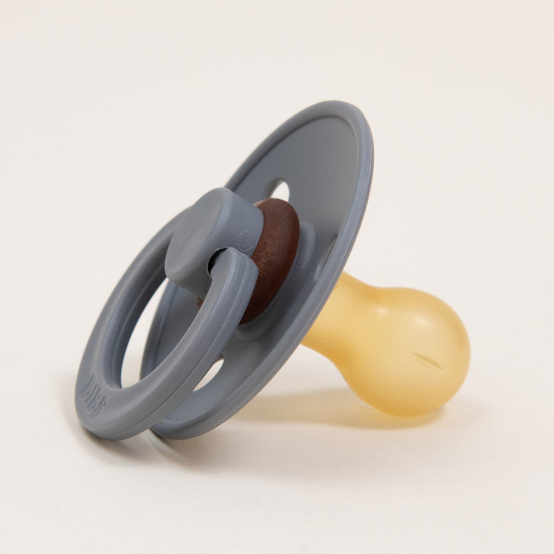 A close-up image of a Bibs Pacifier in Smoke color with a natural gray and brown handle and a yellow nipple, on a light beige background.