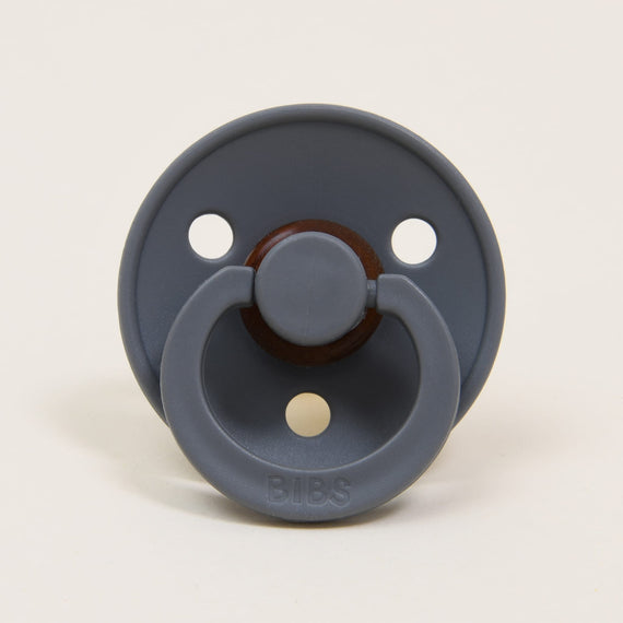 A Bibs Pacifier in Smoke with a round button and "bibs" logo, set against a light beige background, perfect as an upscale baby gift.